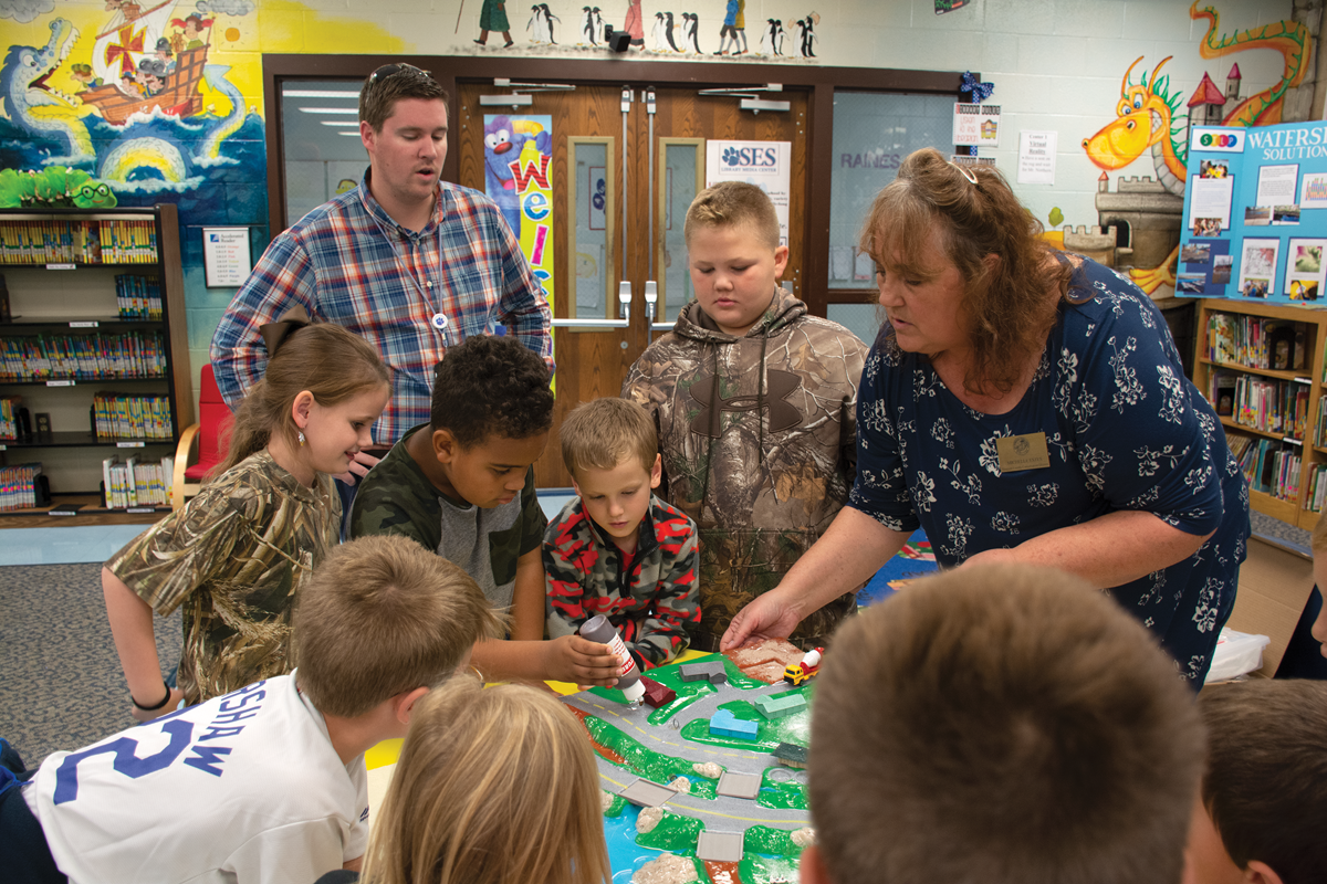Michelle Estes, right, district operation manager for the Simpson County Conservation District, shows Simpson Elementary student Tacarious Savage where to squeeze ‘oil’ onto the model of a watershed as other students look on. 